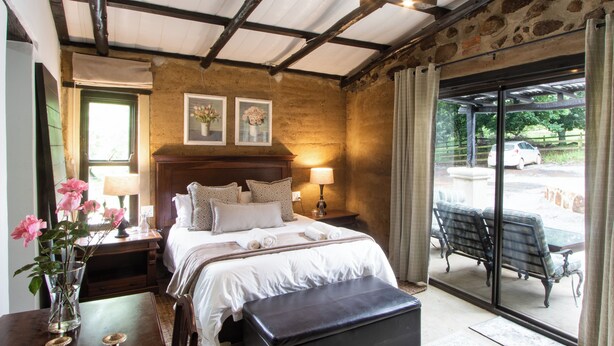 A bedroom with a large bed, bedside tables, a nightstand, a bench, and a view of the outside. - Lake Villa Room 15 (Swartberg)