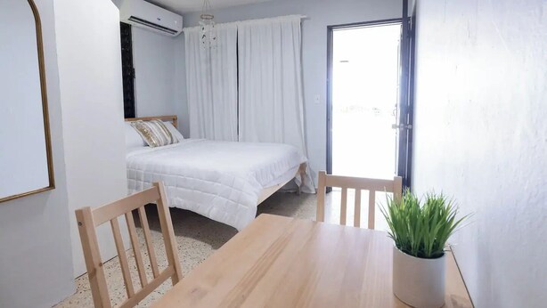 A bedroom with a bed, a wooden table, a potted plant, and a sliding glass door leading to a balcony. - Cozy Studio Type Apartment across Jobos Beach, Isabela. WIFI & A/C. (Isabela)
