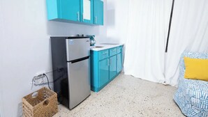 A compact kitchen with a refrigerator, a sink, and teal cabinets. There is a wicker basket on the floor and a bed with a patterned bedspread and a yellow pillow. - Cozy Studio Type Apartment across Jobos Beach, Isabela. WIFI & A/C. (Isabela)