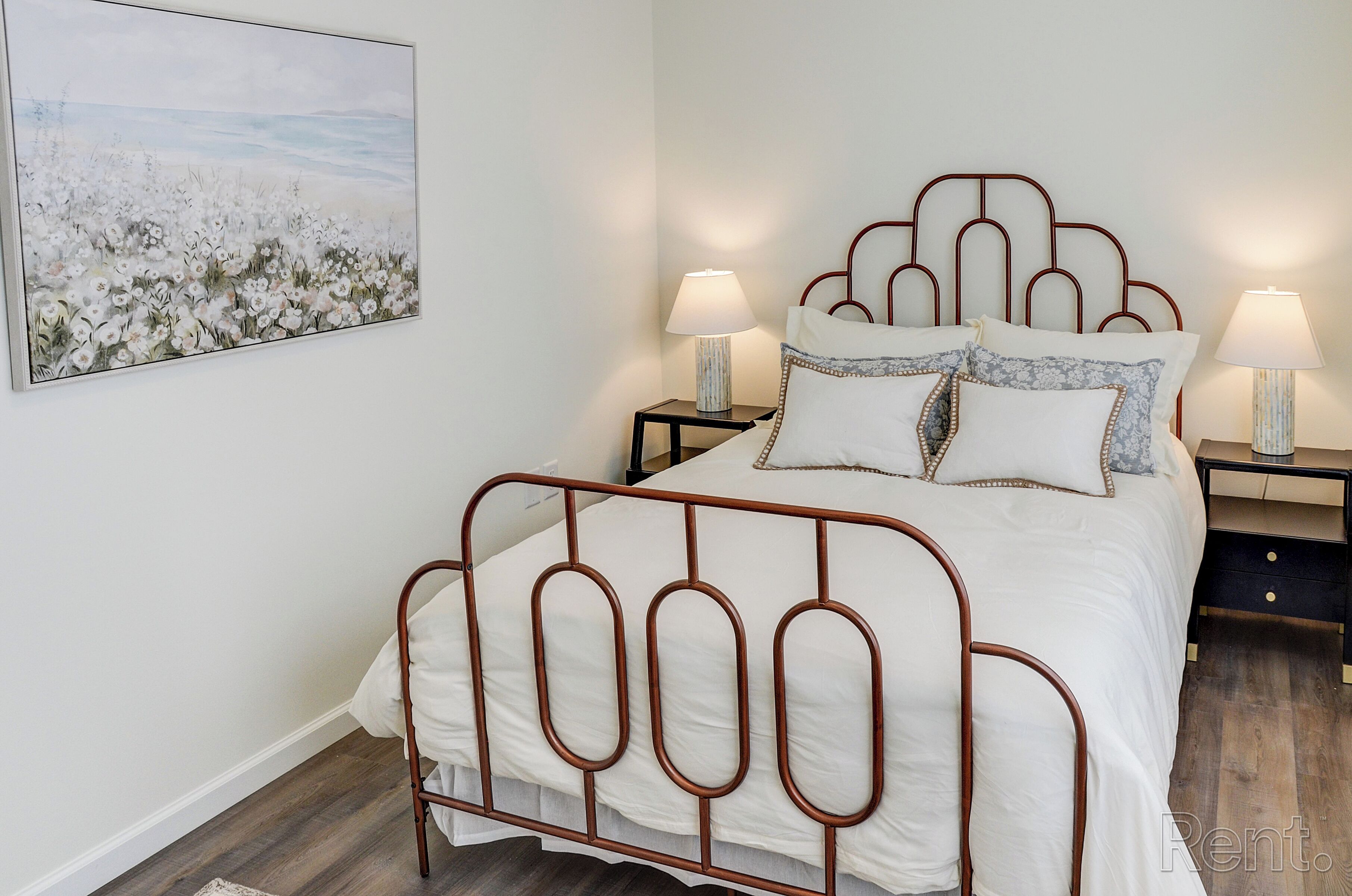 A neatly made bed with a metal frame, two bedside tables with lamps, and a large painting on the wall.
