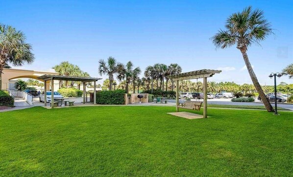 A well-maintained outdoor area with a grassy lawn, a pergola, and palm trees. - Beach Colony East 14D (Pensacola)
