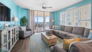 Un salon comprenant un canapé, des fauteuils, une table basse et un téléviseur. Il y a un balcon avec vue sur l’océan.