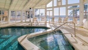 Une piscine intérieure à l’eau claire, entourée de chaises longues et protégée par une paroi de verre offrant une vue sur la plage.