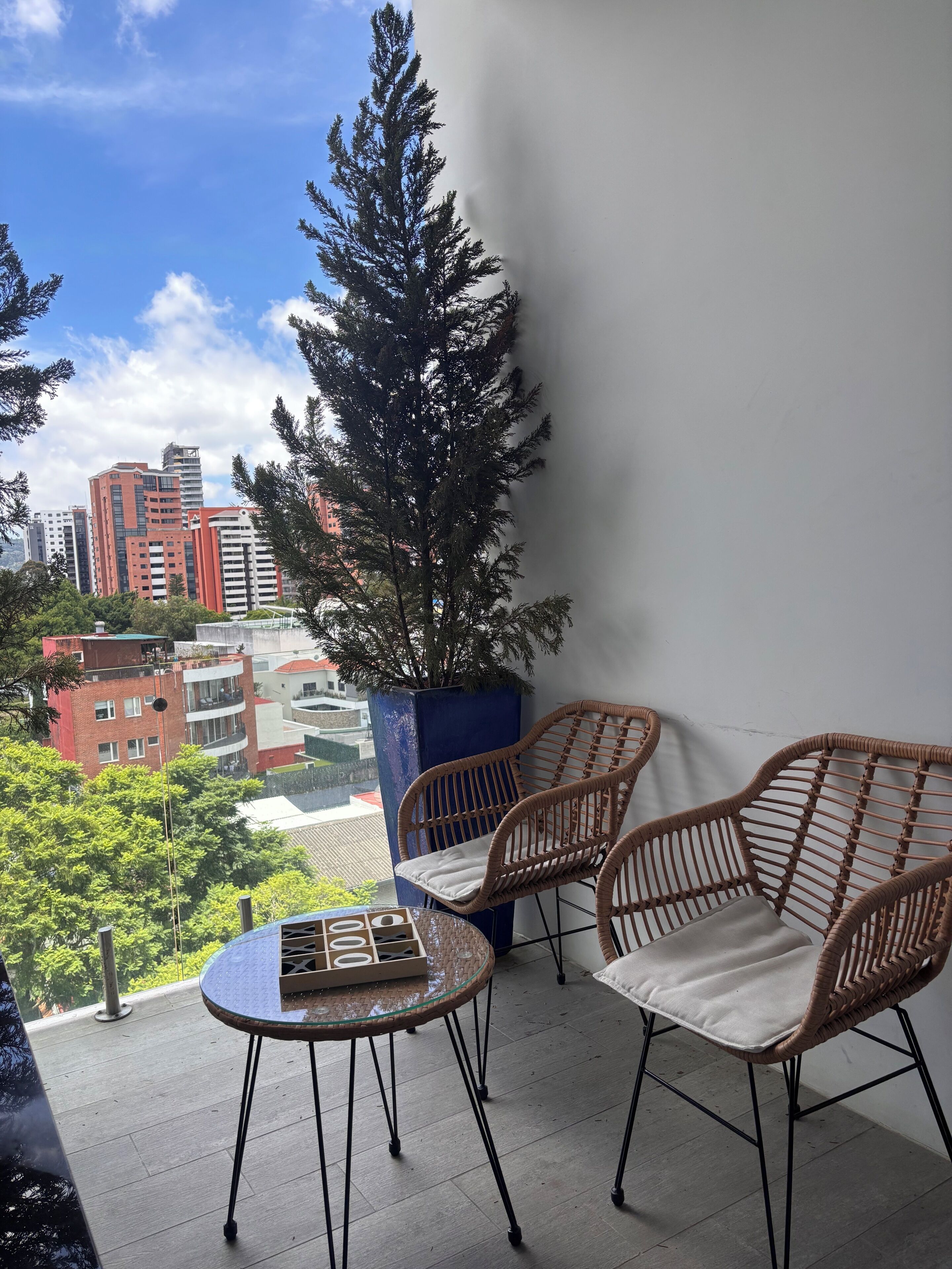 Un balcon avec des fauteuils en osier et une table en verre, d’où l’on peut admirer un panorama urbain avec ses bâtiments et ses arbres.