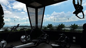 Un gimnasio al aire libre con bicicletas estáticas y una máquina de remo, con vistas a un paisaje urbano y montañas.