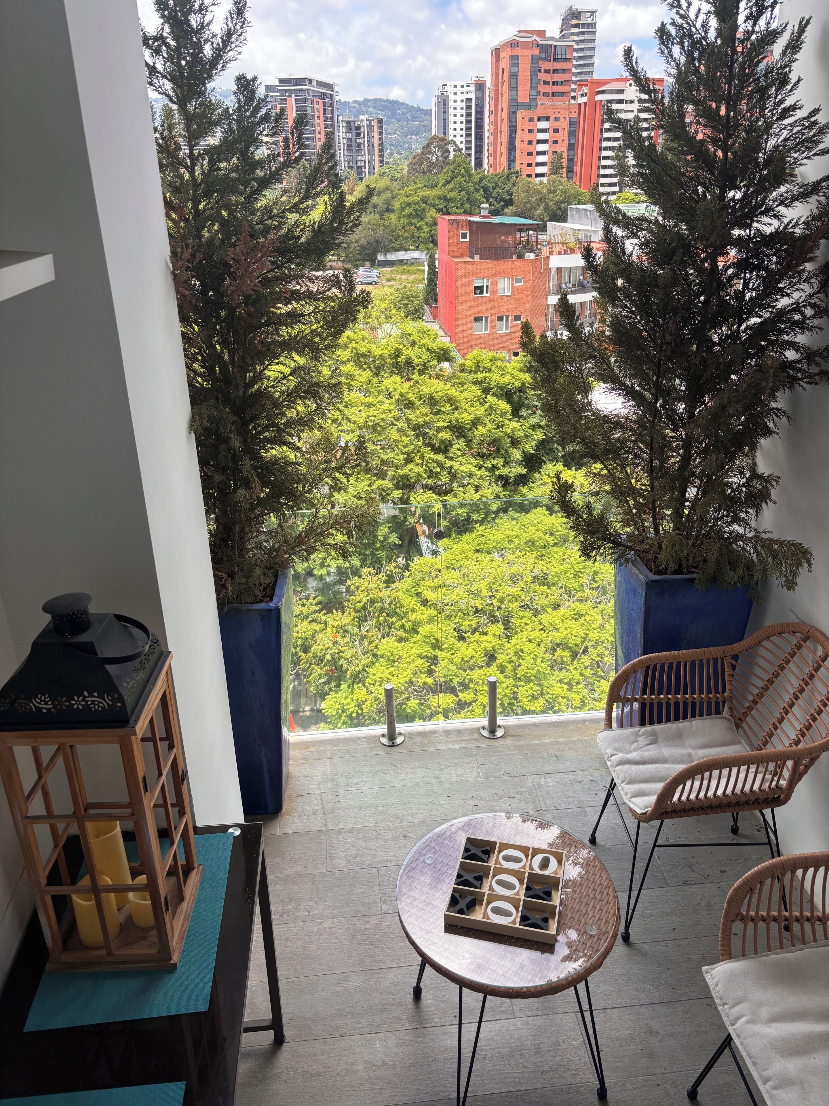 Un balcon donnant sur un paysage urbain, deux plantes en pot, des chaises en osier, une petite table avec un jeu de société et une lanterne en bois.
