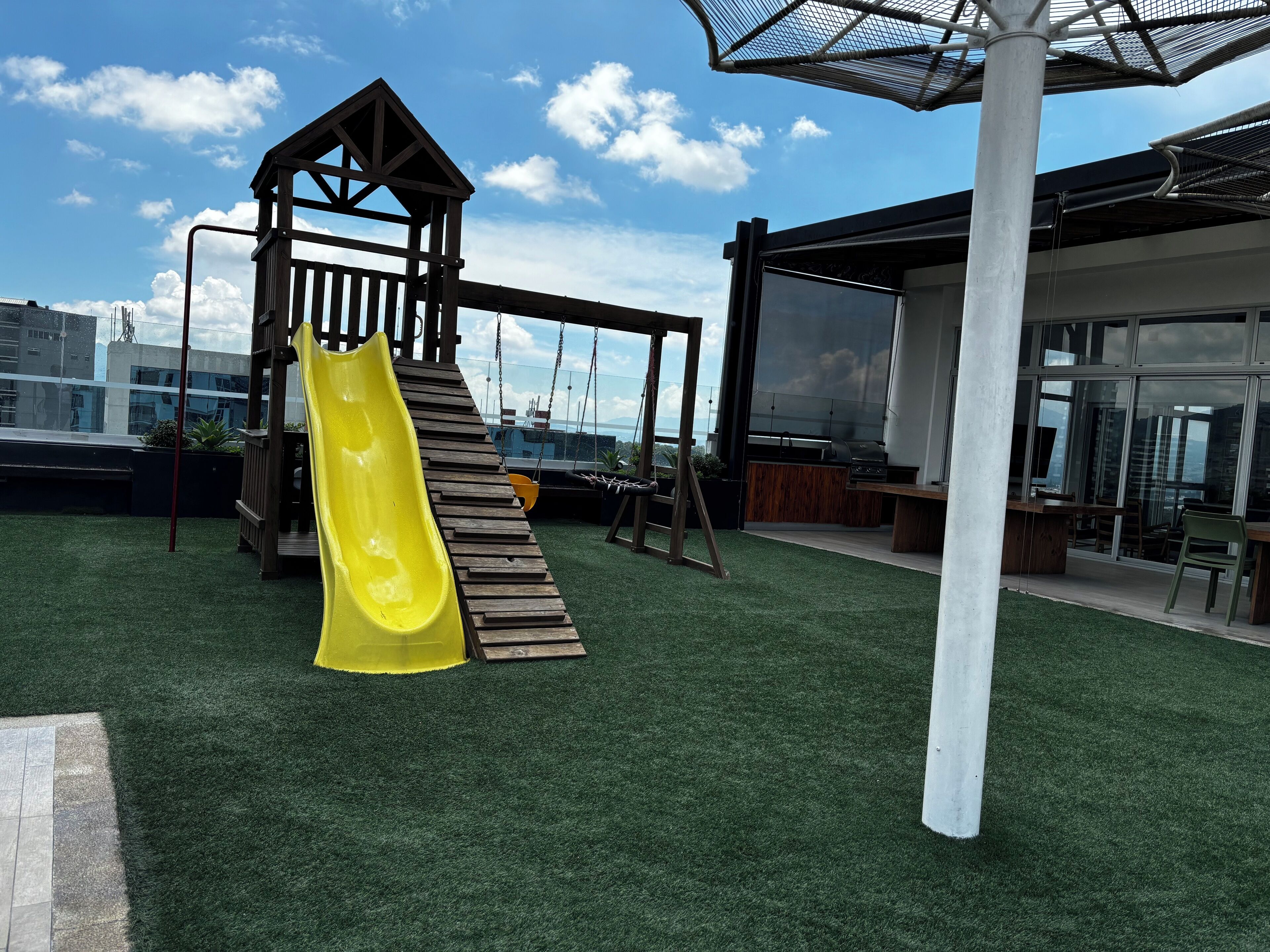 Un terrain de jeux avec un toboggan jaune et une structure de jeu en bois, installé sur une pelouse artificielle, avec en arrière-plan une vue sur des bâtiments.
