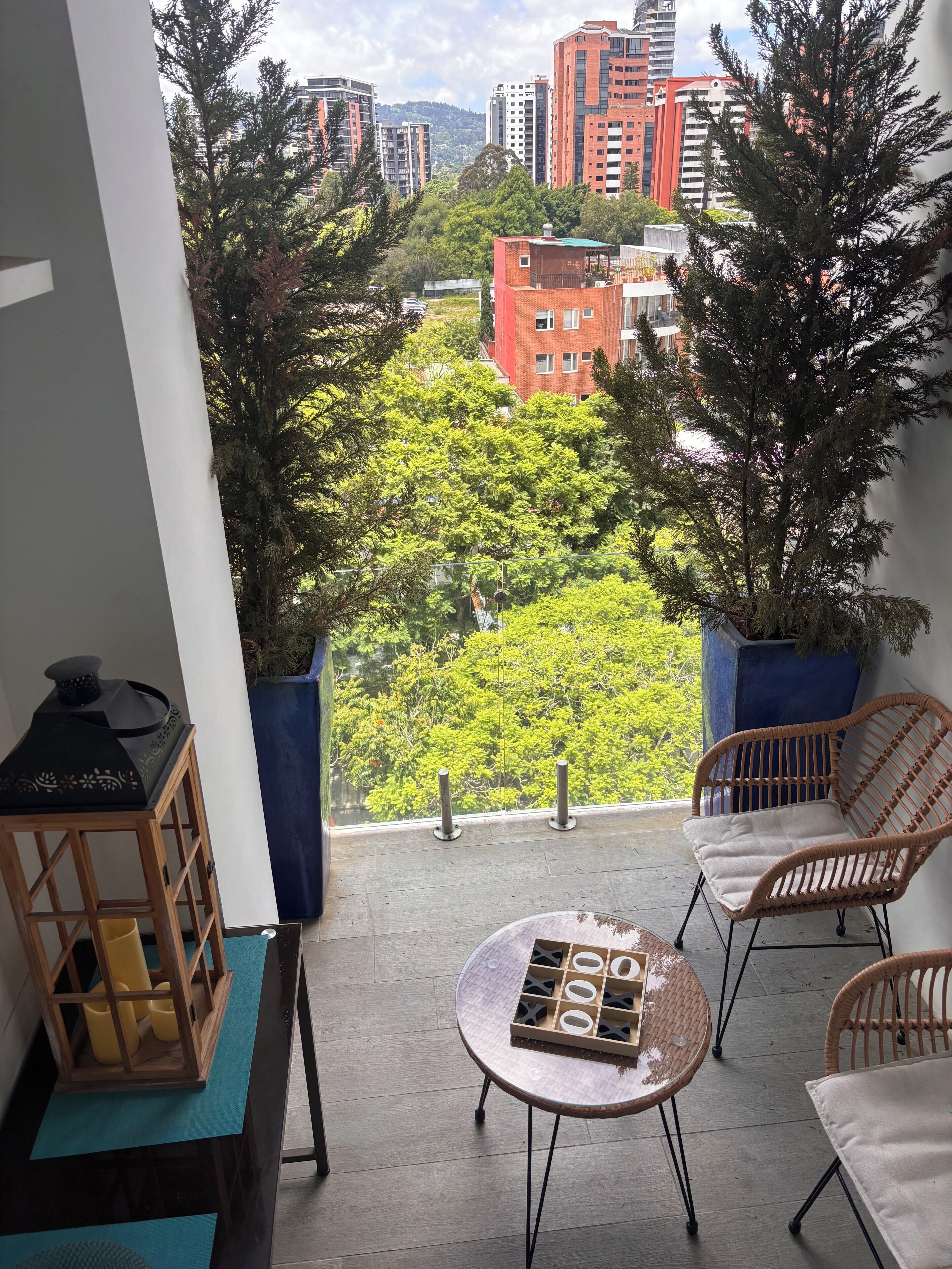 A balcony with a view of a cityscape, two potted plants, a wicker chair, a small table with a board game, and a lantern.