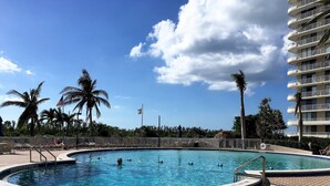 Une piscine avec une terrasse carrelée, des palmiers et un immeuble à plusieurs étages en arrière-plan.