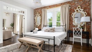 Un dormitorio con pared de ladrillo, una cama grande con ropa de cama blanca, un banco y una mesita de noche con lámpara.