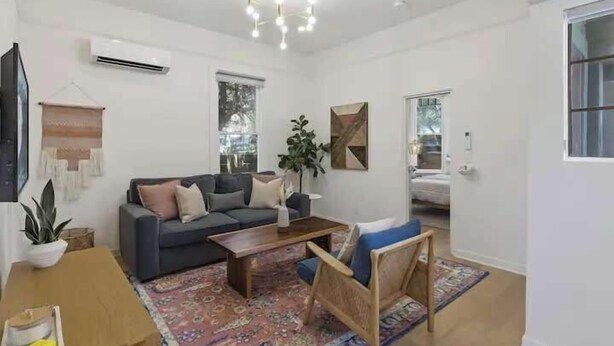 Ein modernes Wohnzimmer mit einer Couch, einem Couchtisch und einem Sessel. Es gibt ein großes Fenster, eine Klimaanlage und eine Zierpflanze.