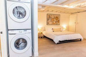 A bedroom with a bed, a nightstand, a wall-mounted artwork, and a washing machine.