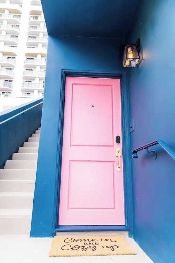 A pink door with a doormat that reads 'Come in & Cozy up' in a building with blue walls and a staircase. - Little Italy Studio: Downtown, Free Parking & More! (San Diego)