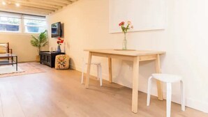 A modern living room with a wooden table, two white stools, a TV mounted on the wall, and a vase with flowers on the table.