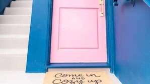 A pink door with a doormat that reads "Come in & Cozy up".