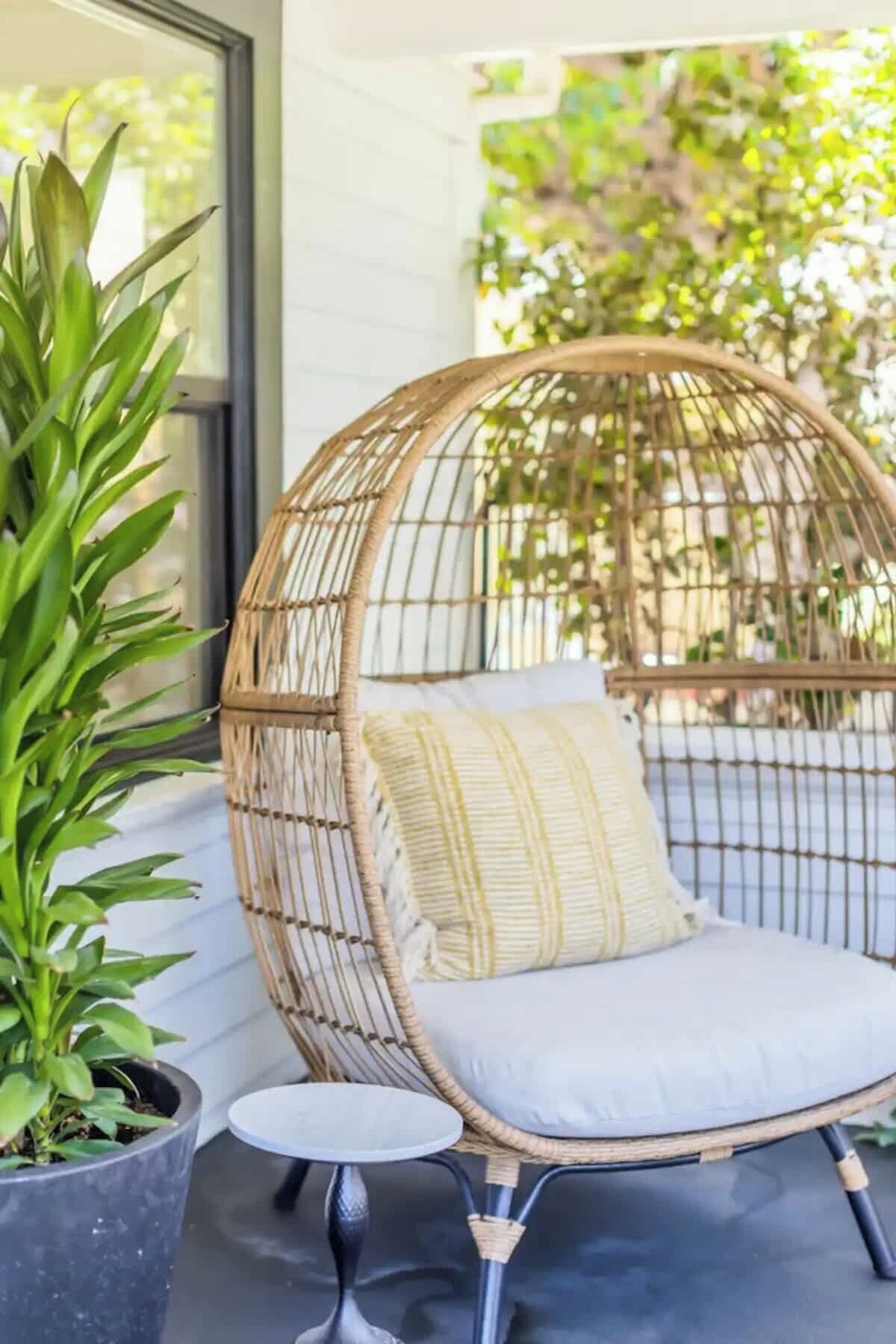 Ein Korbstuhl mit Kissen, ein Blumentopf und ein kleiner runder Tisch auf einer Veranda.