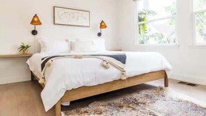 A bedroom with a wooden bed, white bedding, two wall-mounted lamps, a framed picture, a window with a view of greenery, and a small plant on a bedside table. - Bright 4BR Little Italy Home: Free Parking & Sleeps 9! (San Diego)