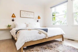 A bedroom with a wooden bed, white bedding, two wall-mounted lamps, a small plant, and a window with a view of greenery.
