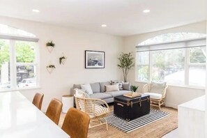 A modern living room with a sofa, coffee table, and chairs. There is a dining table with chairs, a large window, and a plant on the wall.