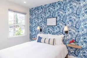 A bedroom with a bed, a window, a wall with a leaf pattern, two wall-mounted lamps, and a framed item on the wall.