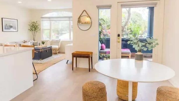 A modern living room with a round dining table, a sofa, and a coffee table. There is a window with a view of greenery outside. - Bright 4BR Little Italy Home: Free Parking & Sleeps 9! (San Diego)