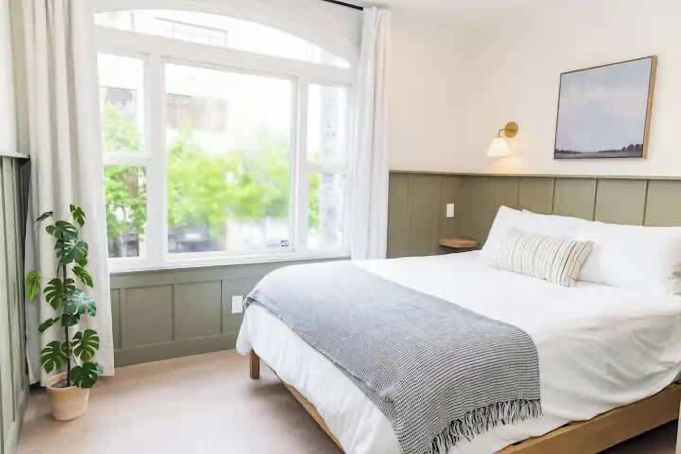 Ein Schlafzimmer mit Bett, Fenster, Pflanze und einer Wandleuchte.