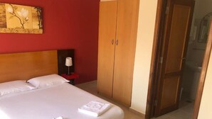 A neatly made bed with white linens and a folded towel, a wooden wardrobe, and a nightstand with a lamp. - Tarrafal Residence (Tarrafal)
