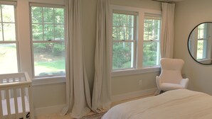 A bedroom with a crib, a chair, and a bed. There are windows with curtains and a view of greenery outside. - Corkscrew Cape - Family Friendly Upstate NY Escape (Barryville)