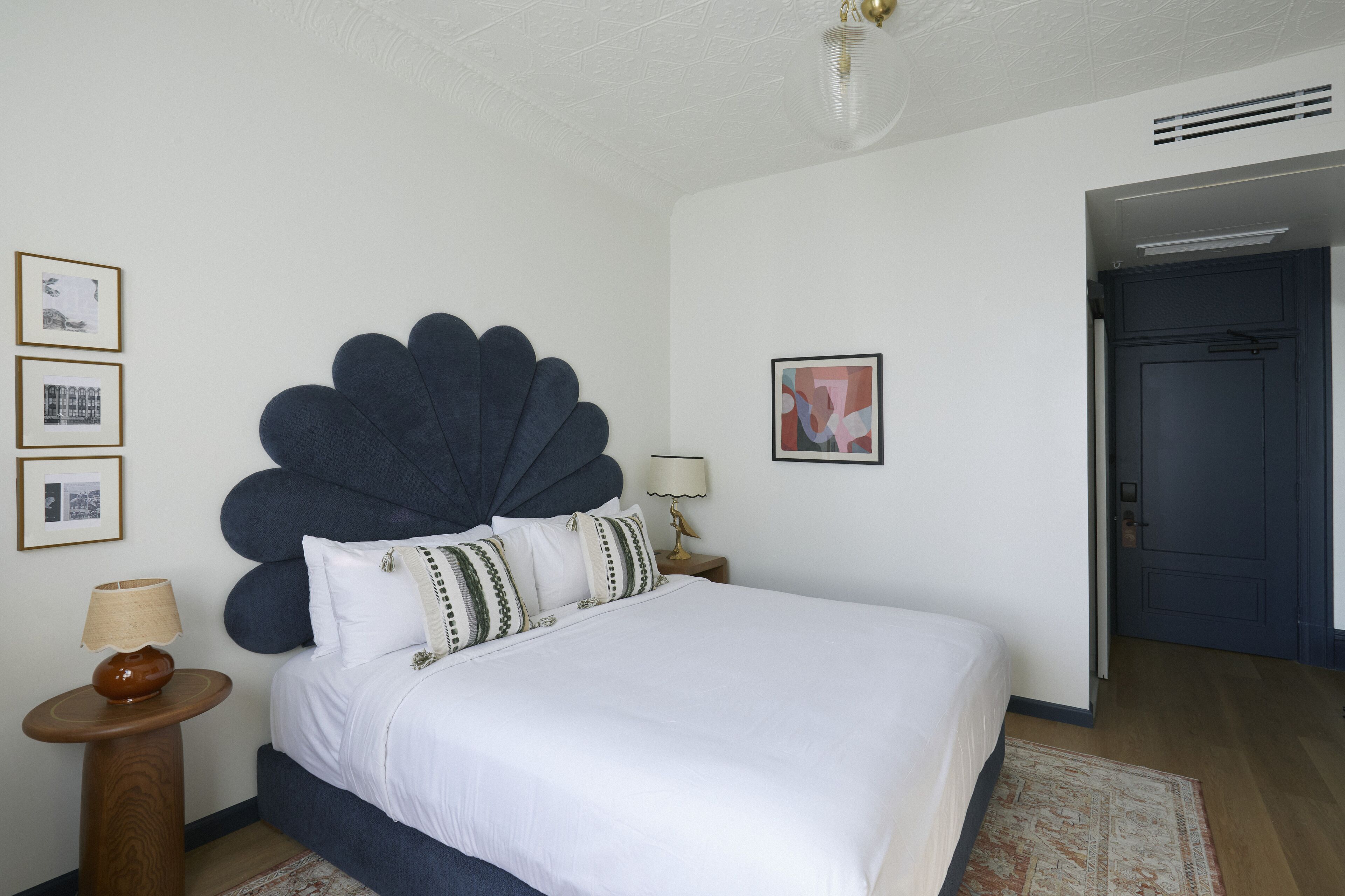 A bedroom with a large bed, a round wooden nightstand, a wall-mounted lamp, framed pictures, and a dark door.