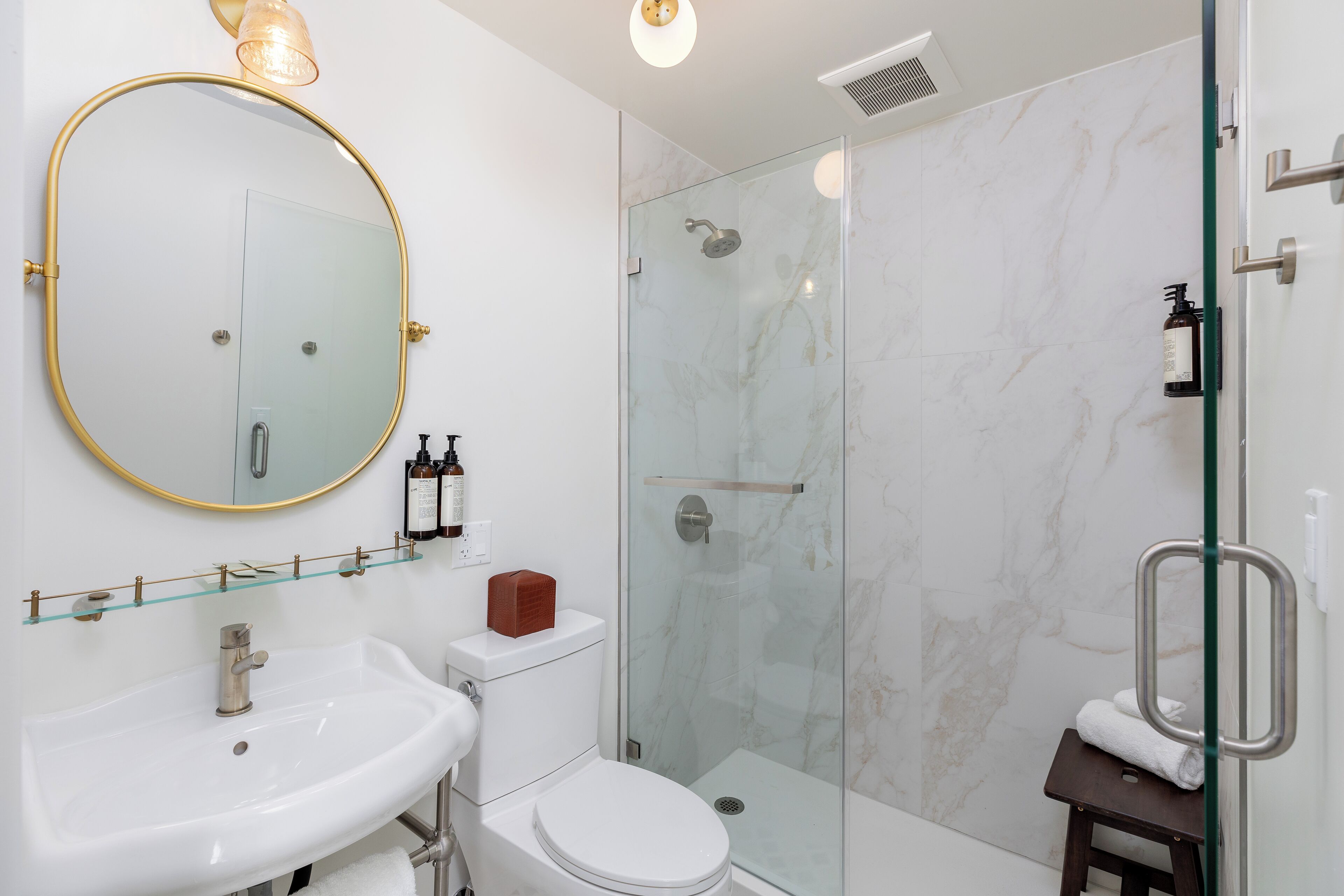 Un bagno moderno con doccia vetrata, un lavandino bianco e uno specchio tondo con cornice dorata.
