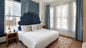 Une chambre moderne avec un grand lit, une tête de lit décorative, une table de chevet et une vue sur la ville grâce à de grandes fenêtres.
