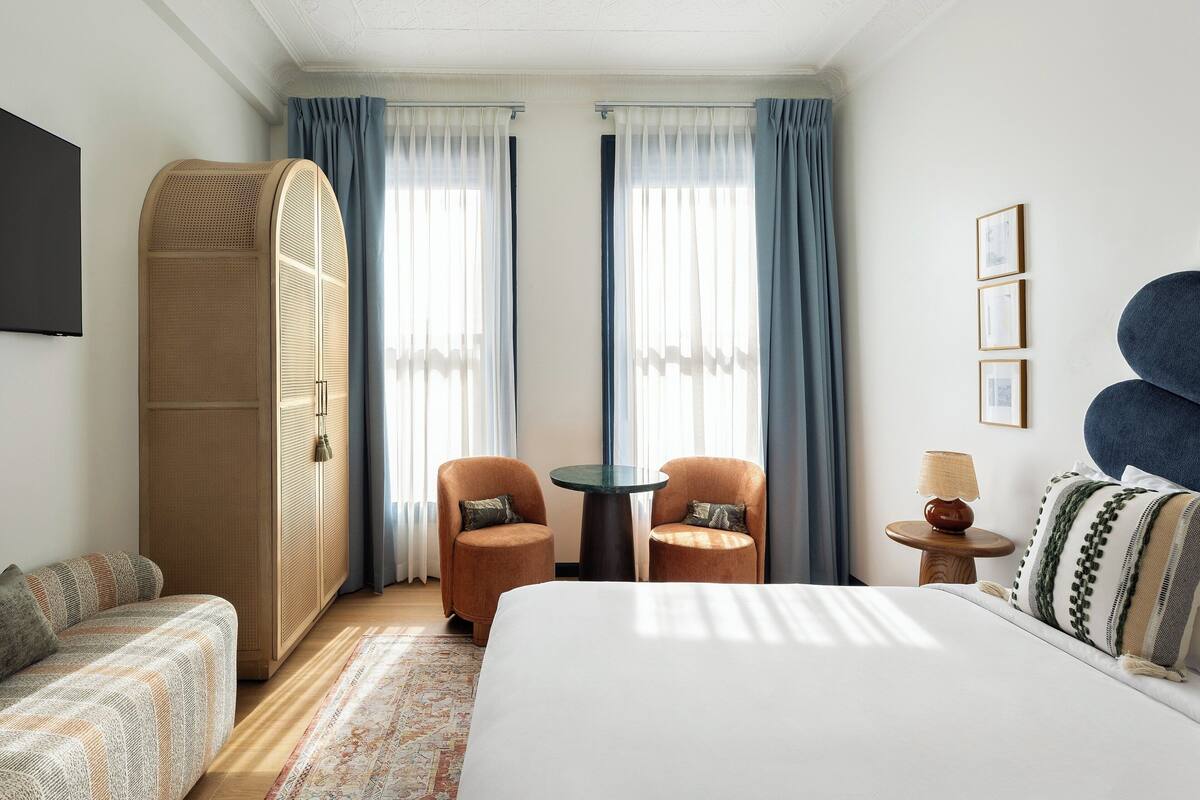 A hotel room with a bed, two armchairs, a small table, a lamp, and a television.
