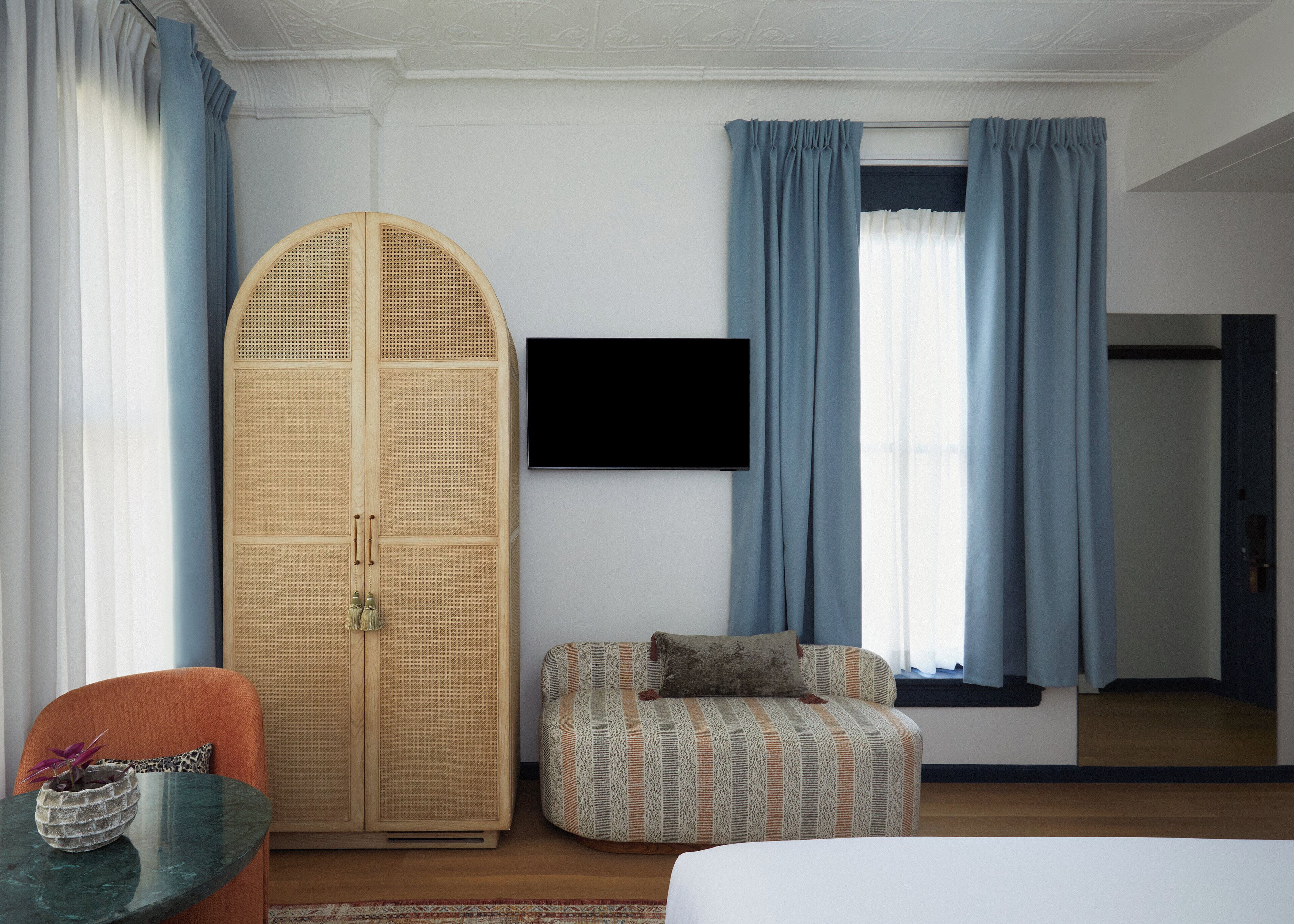 A hotel room with a bed, a television mounted on the wall, a wardrobe, a seating area with a patterned sofa, and a small table with a plant.