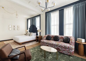 Ein Hotelzimmer mit Bett, Sofa, Couchtisch, Sessel, Lampe und Lautsprecher.