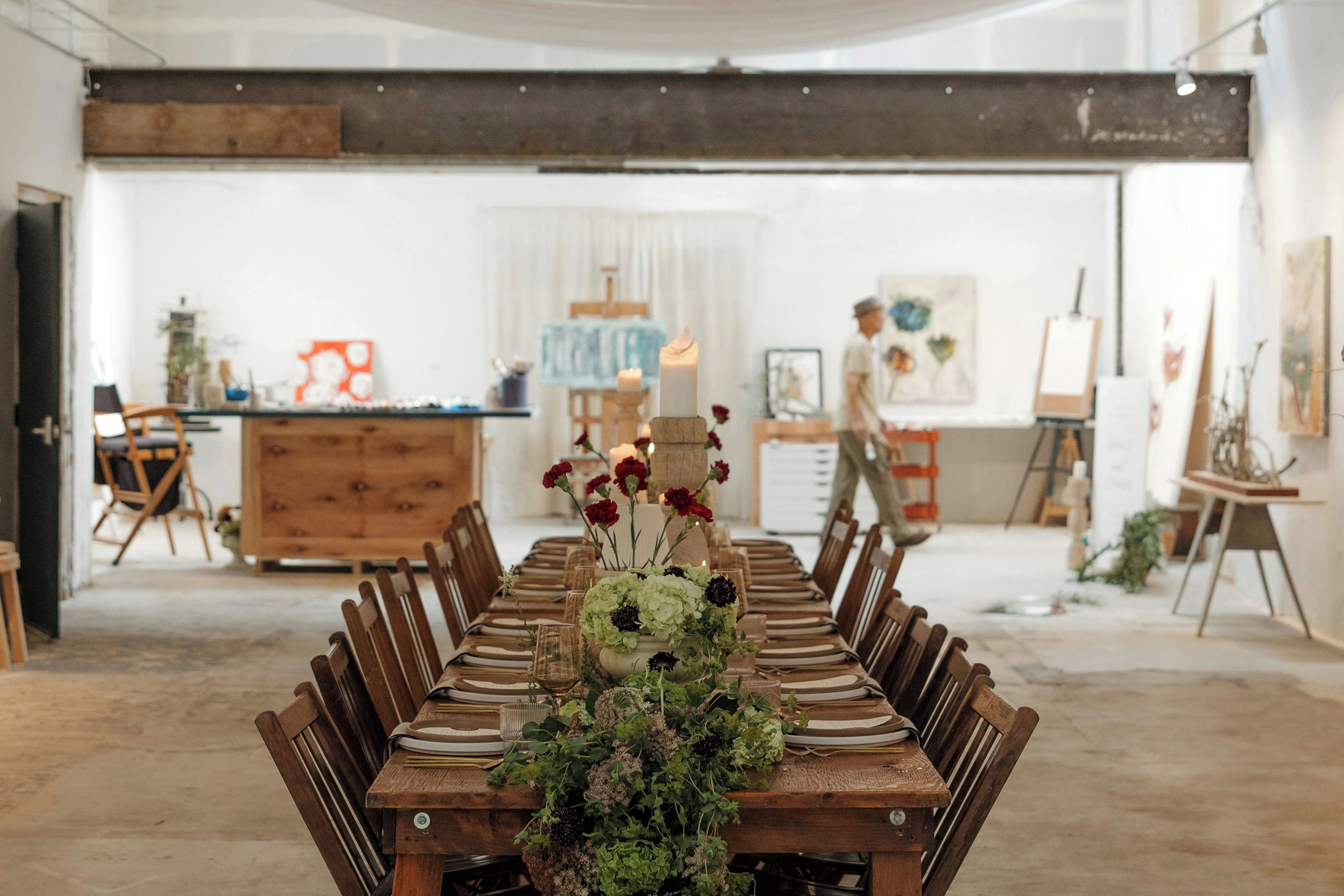 Una larga mesa de comedor lista para una comida, con sillas a ambos lados, un centro de mesa con flores y una habitación con arte en las paredes y un ventanal grande.