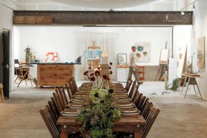 Een lange eettafel gedekt voor een maaltijd, met stoelen aan beide zijden, een bloemenarrangement als middelpunt en een kamer met kunstwerken aan de muur en een groot raam.