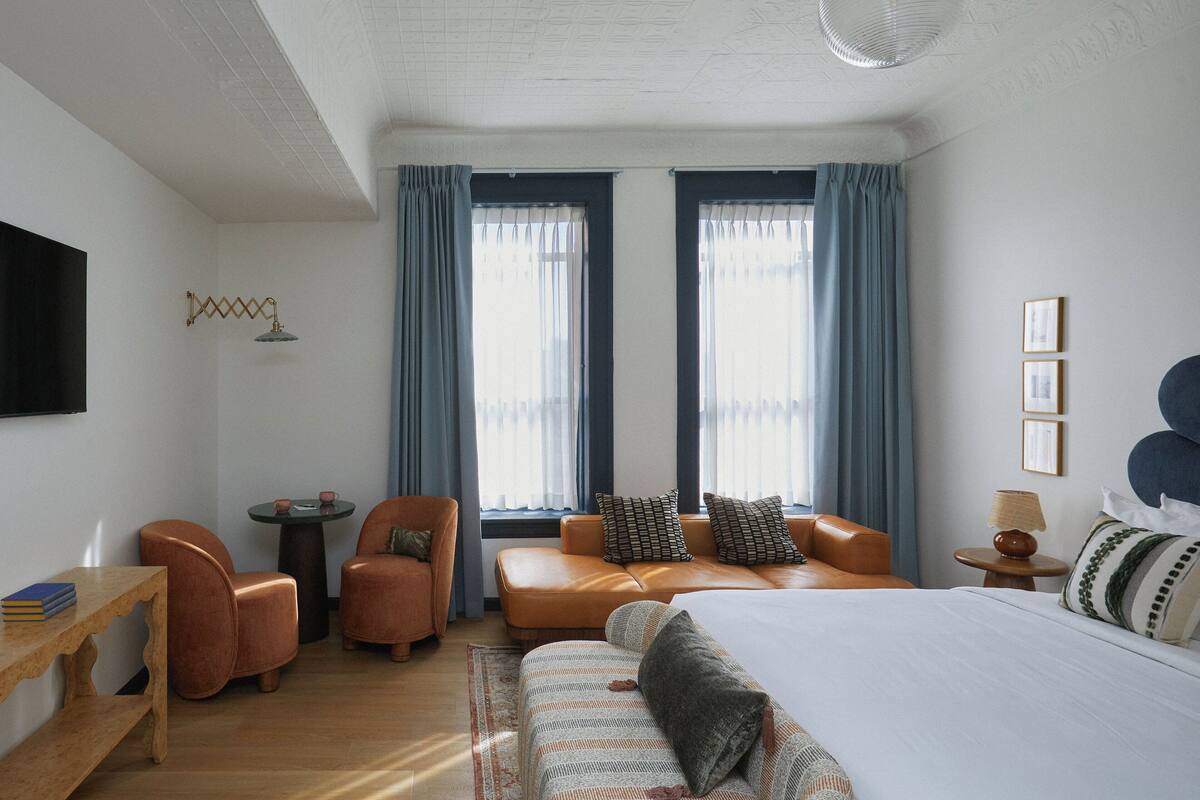 A modern hotel room with a bed, two windows with curtains, a sofa, a chair, a small table, and a lamp.
