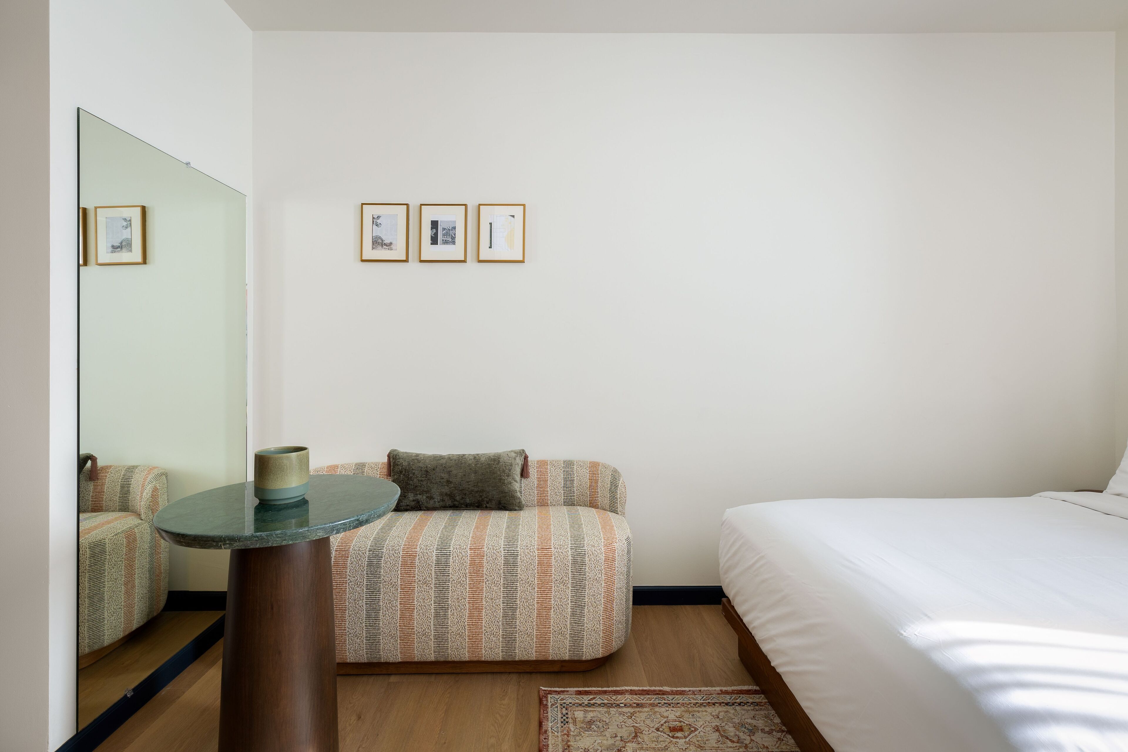 A hotel room with a bed, a sofa, a small table, and a mirror. There are three framed pictures on the wall.