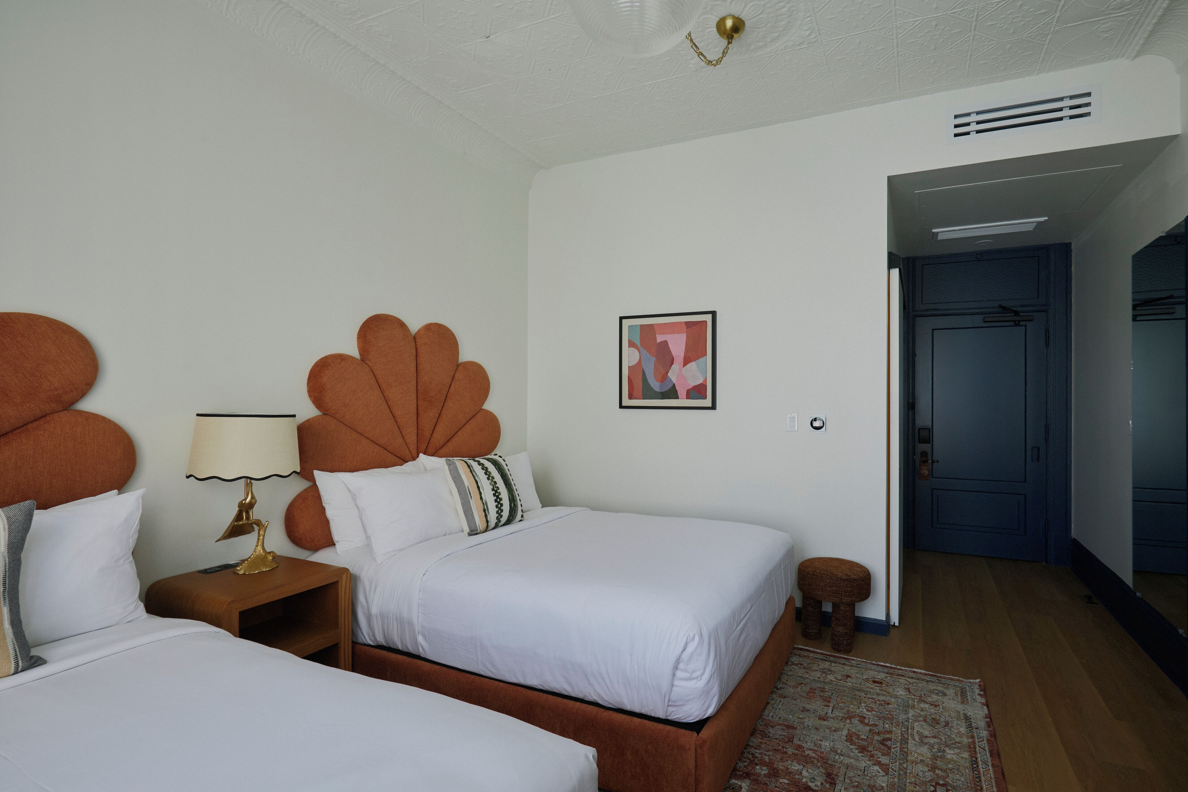 Ein Hotelzimmer mit zwei Betten, Holzboden, einem gemusterten Teppich, einer Wandlampe und einer Tür zu einem weiteren Raum.