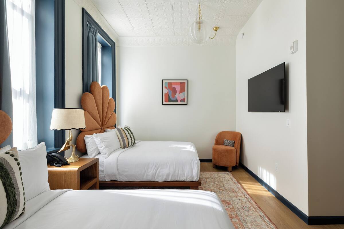 A hotel room with a bed, bedside table, lamp, chair, TV, artwork, and a patterned rug.