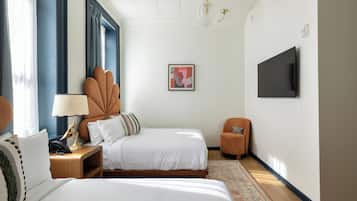 A hotel room with a bed, bedside table, lamp, chair, TV, artwork, and a patterned rug.