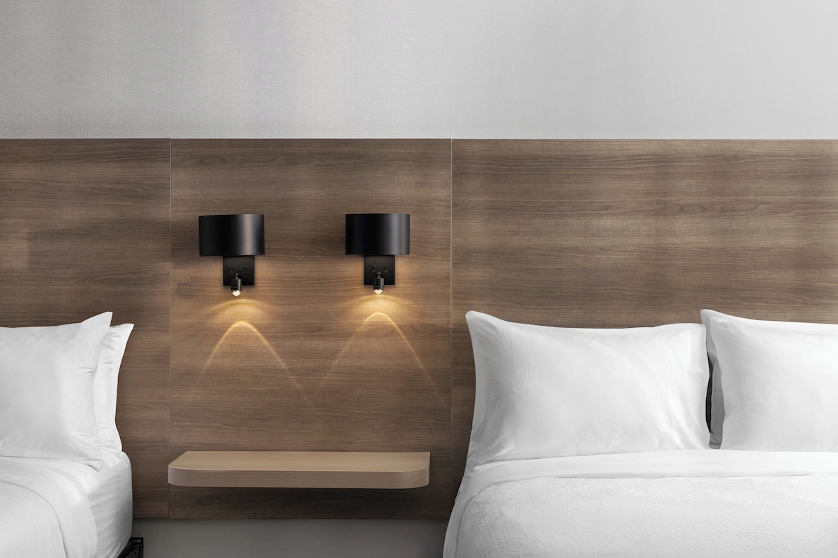 Una habitación de hotel con cabecera de madera, dos lámparas de noche, mesitas de noche y ropa de cama blanca.