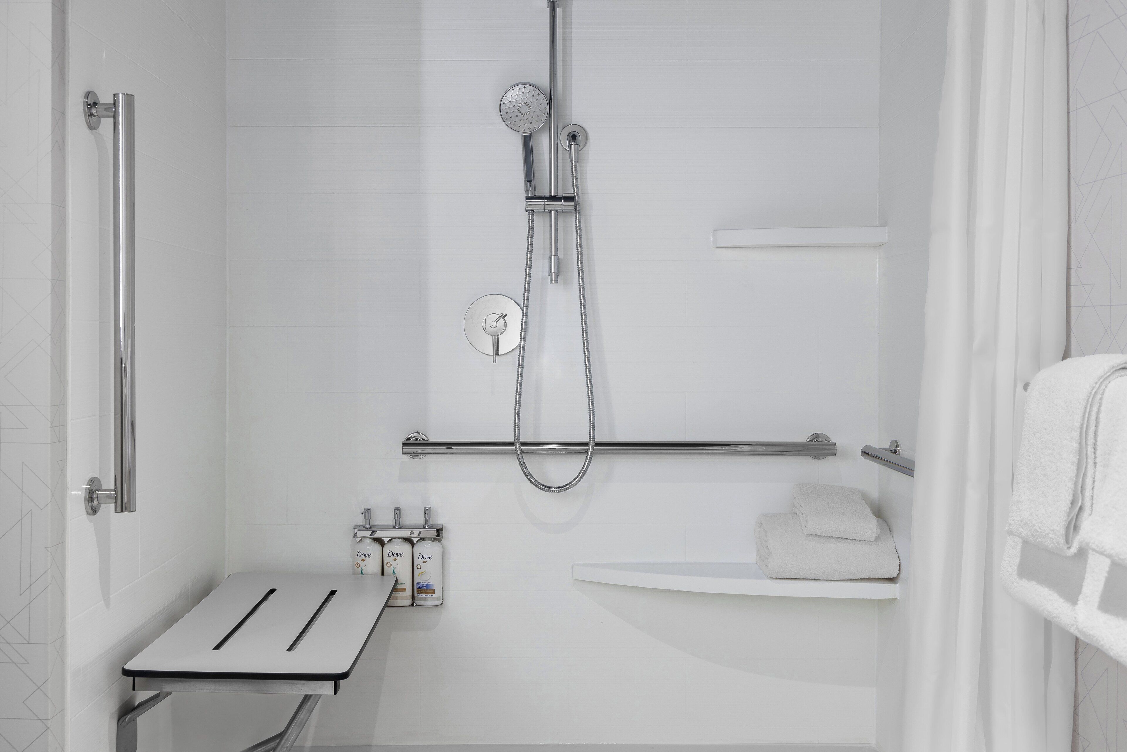A modern shower with a handheld showerhead and a wall-mounted faucet. There is a towel rack with folded towels and a small shelf with toiletries.