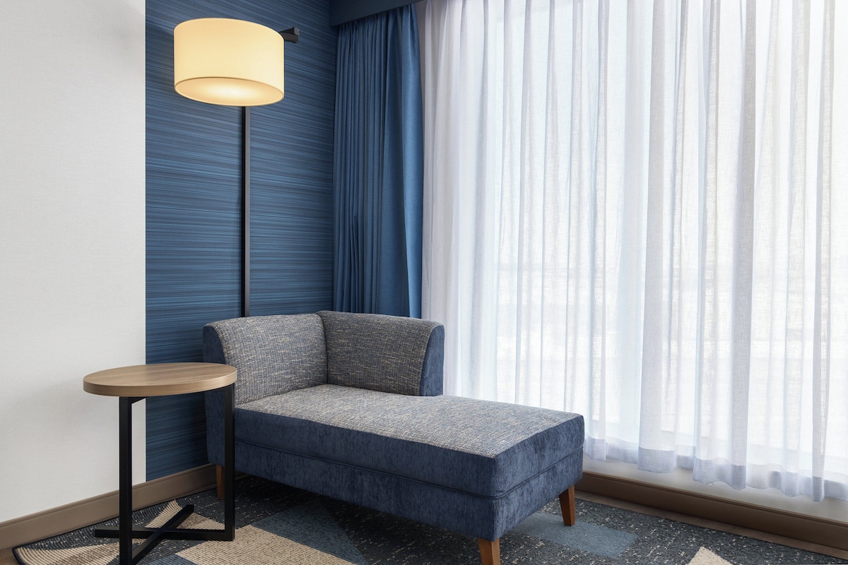 Habitación de esquina con sofá azul, una pequeña mesa redonda, una lámpara de pie y cortinas traslúcidas.