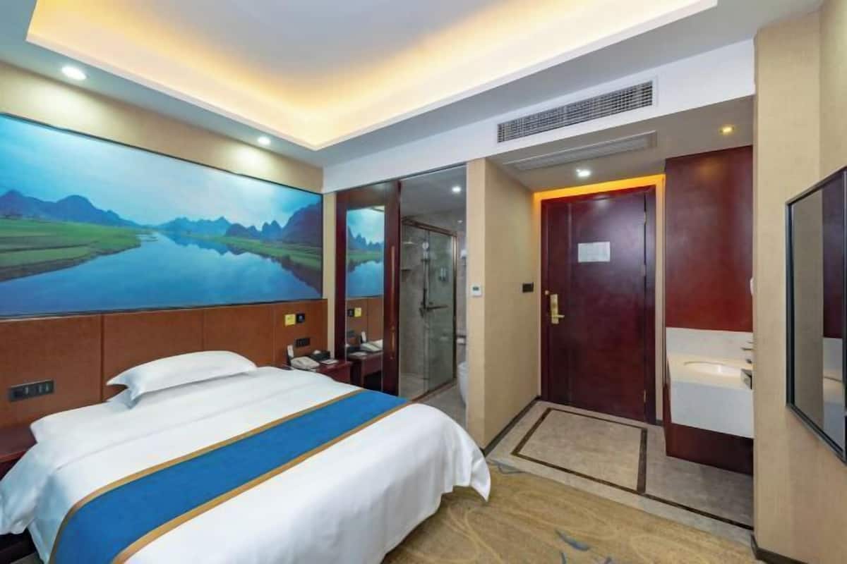 A hotel room with a bed, a painting, a bathroom, and a shower area.