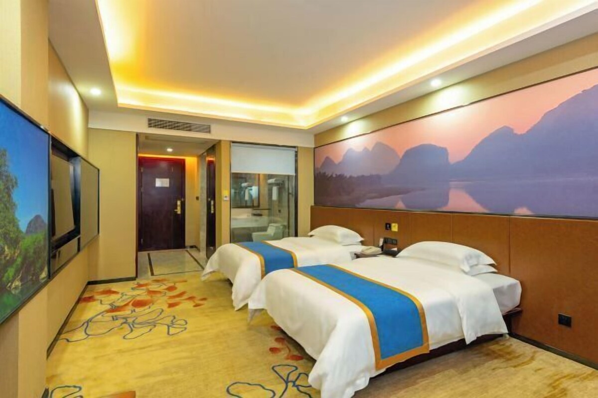 A hotel room with two beds, a large window with a mountain view, and a carpet with a floral pattern.