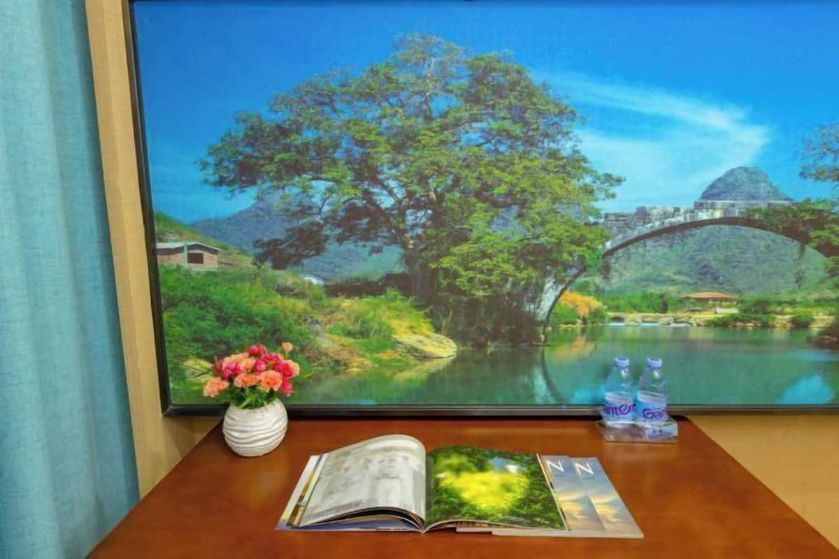 A room with a large window displaying a scenic view of a river, trees, and a bridge. There is a book, a vase with flowers, and two water bottles on the table.