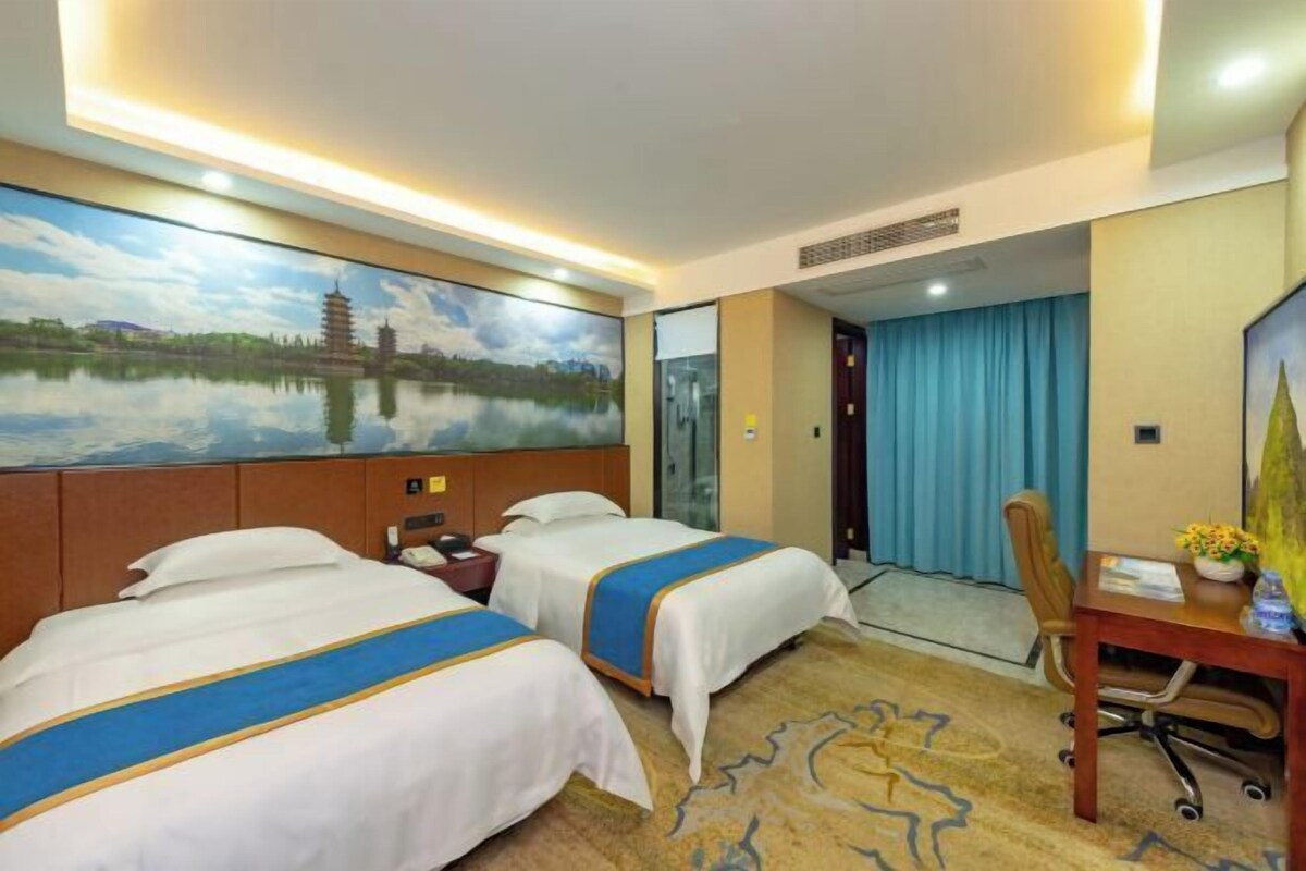 A hotel room with two beds, a desk, a chair, a painting of a pagoda, and a view of a building through a glass door.