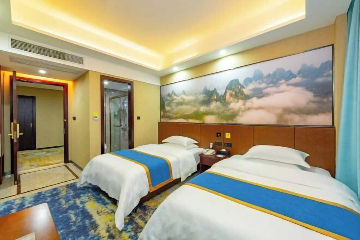 A hotel room with two beds, a large painting of mountains, and a carpeted floor.