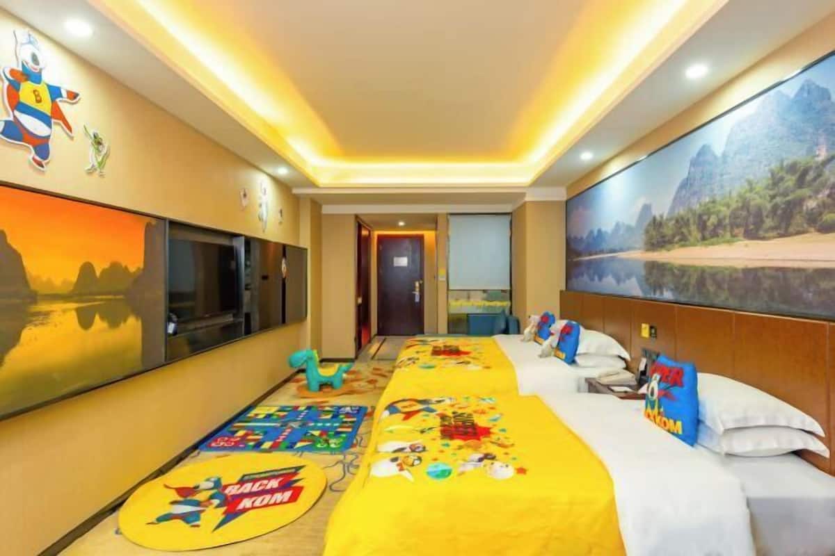 A hotel room with a bed, a large TV, a painting, and a children's play mat.